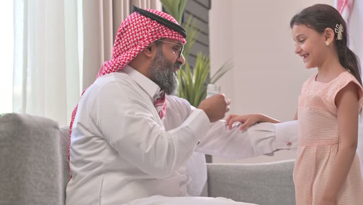 التجمعات والزيارات الاسرية في الأعياد والمناسبات الاسلامية, رجل عربي خليجي سعودي يرتدي الثوب التقليدي والشماغ يقوم  بتقديم العيديه لابنته، صلة الرحم والترابط الاسري, فرحة وبهجة الابناء في ايام العيد السعيد