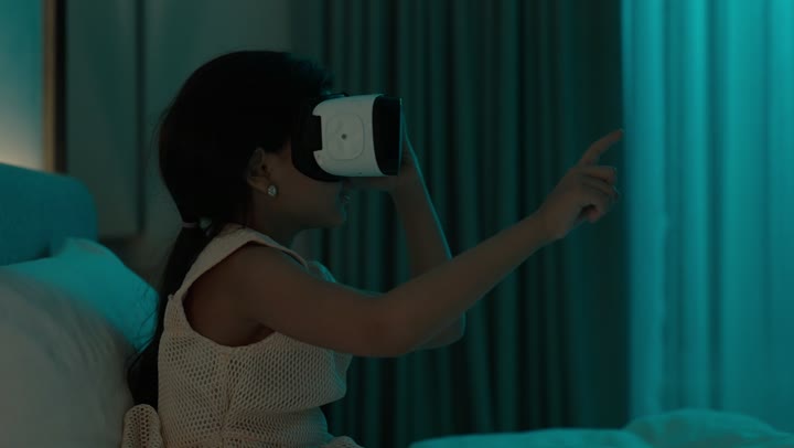 تعابير الاندماج والتركيز، فتاة عربية خليجية سعودية تجلس على السرير تقوم بمشاهدة الأفلام عبر نظارات الواقع الافتراضي ثلاثية الأبعاد، محاكاة العالم الخيالي والتفاعل معه محاكاة العالم الخيالي، استخدام جهاز تقني حديث ومتطور