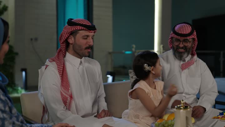 صلة الرحم والترابط الاسري, فرحة وبهجة الابناء في ايام العيد السعيد, التجمعات والزيارات الاسرية في الأعياد والمناسبات الاسلامية, رجل عربي خليجي سعودي يرتدي الزي التقليدي يجلس مع أحفاده بإيماءات الفرح والسرور، ضيافة وحلوى العيد، حسن استقبال وإكرام الضيف
