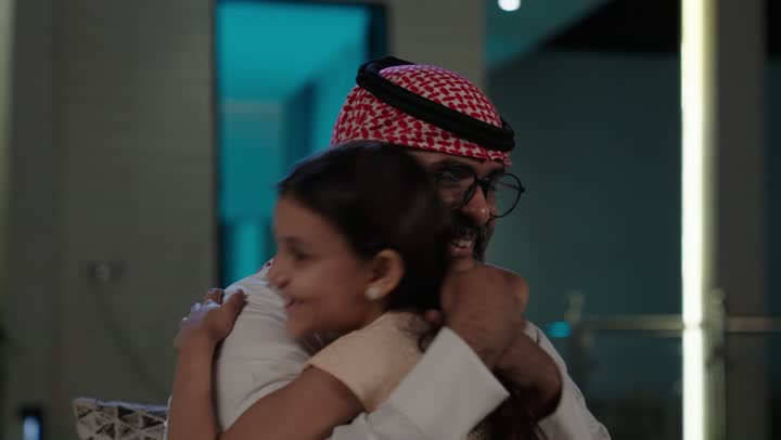 مفهوم الحب والحنان، دور الأب في حياه الأبناء، تصوير مقرب لأب عربي خليجي سعودي يرتدي الثوب التقليدي والشماغ يجلس على كرسي يحتضن ابنته بين ذراعيه، قضاء وقت ممتع ومسلي، ايماءات السعادة والسرور،  اجواء اسرية مليئة بالفرح