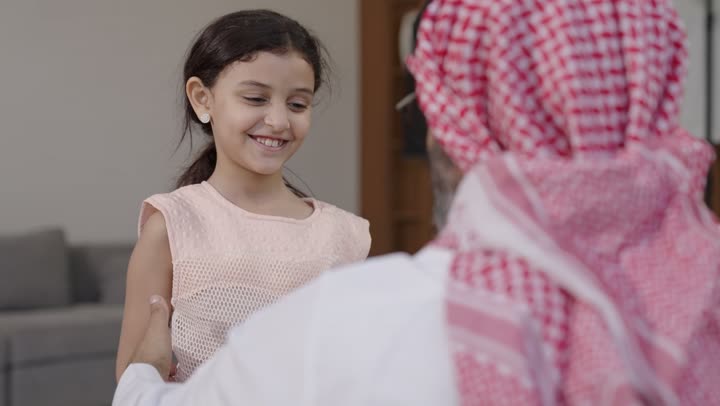 مفهوم الحب والحنان، دور الأب في حياه الأبناء، تصوير مقرب لأب عربي خليجي سعودي يرتدي الثوب التقليدي والشماغ يجلس على كرسي يحتضن ابنته بين ذراعيه، قضاء وقت ممتع ومسلي، ايماءات السعادة والسرور،  اجواء اسرية مليئة بالفرح