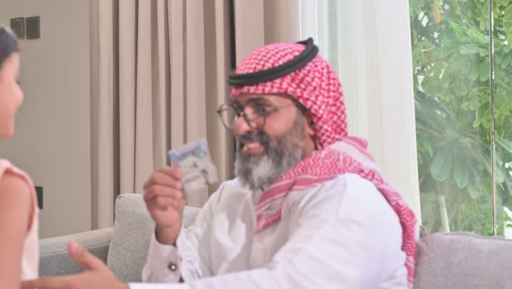 تقديم العيدية في العيد، مفهوم الحب والحنان، دور الأب في حياه الأبناء، تصوير مقرب لأب عربي خليجي سعودي يرتدي الثوب التقليدي والشماغ يجلس على كرسي يحتضن ابنته بين ذراعيه، قضاء وقت ممتع ومسلي، ايماءات السعادة والسرور،  اجواء اسرية مليئة بالفرح