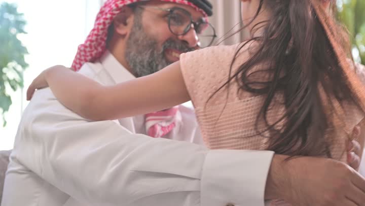 مفهوم الحب والحنان، دور الأب في حياه الأبناء، تصوير مقرب لأب عربي خليجي سعودي يرتدي الثوب التقليدي والشماغ يجلس على كرسي يحتضن ابنته بين ذراعيه، قضاء وقت ممتع ومسلي، ايماءات السعادة والسرور،  اجواء اسرية مليئة بالفرح