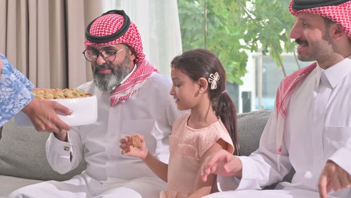 ضيافة وحلوى العيد، حسن استقبال وإكرام الضيف، صلة الرحم والترابط الاسري, فرحة وبهجة الابناء في ايام العيد السعيد, التجمعات والزيارات الاسرية في الأعياد والمناسبات الاسلامية, رجل عربي خليجي سعودي يرتدي الثوب التقليدي يجلس مع أحفاده بإيماءات الفرح والسرور