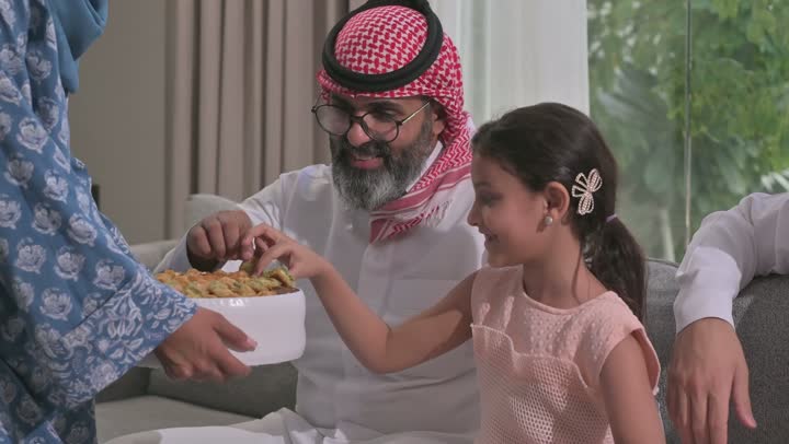 ضيافة وحلوى العيد، حسن استقبال وإكرام الضيف، صلة الرحم والترابط الاسري, فرحة وبهجة الابناء في ايام العيد السعيد, التجمعات والزيارات الاسرية في الأعياد والمناسبات الاسلامية, رجل عربي خليجي سعودي يرتدي الثوب التقليدي يجلس مع أحفاده بإيماءات الفرح والسرور