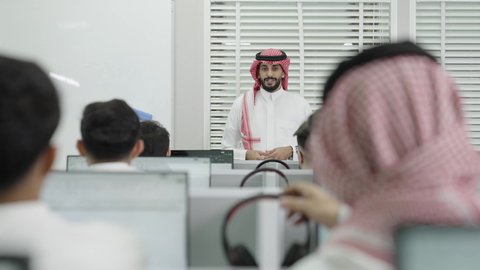 Lectures and educational courses in Saudi Arabia, use of headphones, a classroom equipped with screens and modern technological devices, knowledge and education in Saudi society, holding training courses for students, a speaker and a university professor, an Arab Gulf Saudi wearing a traditional headscarf and thobe, giving a lecture in front of students.