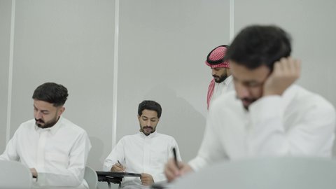 Studying and striving for excellence, workshops and training courses, higher education in the Kingdom of Saudi Arabia, an Arab Gulf Saudi university professor wearing the traditional headscarf and thobe supervising students, university students taking exams, taking notes and writing reports.