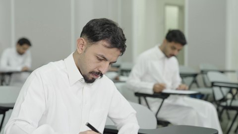 التعليم الجامعي في المملكة العربية السعودية, ورش عمل ودورات تدريبية، التفكير في الإجابة الصحيحة، الدراسة والاجتهاد من اجل التفوق، طالب جامعي عربي خليجي سعودي يرتدي الثوب التقليدي يقوم بتقديم الاختبارات، تدوين وكتابة الملاحظات والتقارير