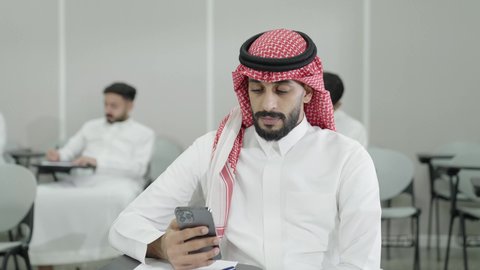 جهاز تقني حديث ومتطور، التعليم الجامعي في المملكة العربية السعودية, ورش عمل ودورات تدريبية، طالب جامعي عربي خليجي سعودي يرتدي الثوب والشماغ التقليدي يقوم باستخدام الهاتف المحمول داخل القاعة الدراسية