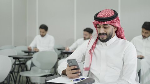 ايماءات السرور والسعادة، جهاز تقني حديث ومتطور، التعليم الجامعي في المملكة العربية السعودية, ورش عمل ودورات تدريبية، طالب جامعي عربي خليجي سعودي يرتدي الثوب والشماغ التقليدي يقوم باستخدام الهاتف المحمول داخل القاعة الدراسية