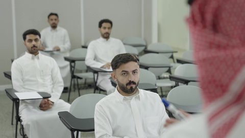 التدريس الفعال في السعودية، التعليم العالي في الجامعات والكليات, دمج التعلم مع التكنولوجيا، تصوير من الخلف لأستاذ جامعي عربي خليجي سعودي يرتدي الثوب التقليدي والشماغ يقوم بشرح المعلومات على الطلاب، جودة التعليم الجامعي في المملكة العربية السعودية