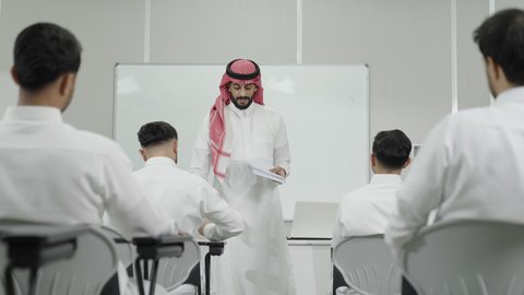 التفاعل داخل الفصل الدراسي، التعليم العالي في الجامعات والكليات الأكاديمية, اختبارات وكتابة التقارير والأبحاث العلمية، أستاذ جامعي عربي خليجي سعودي يرتدي الثوب التقليدي والشماغ يقوم بتوزيع الأوراق على الطلاب، جودة التعليم الجامعي في المملكة العربية السعودية