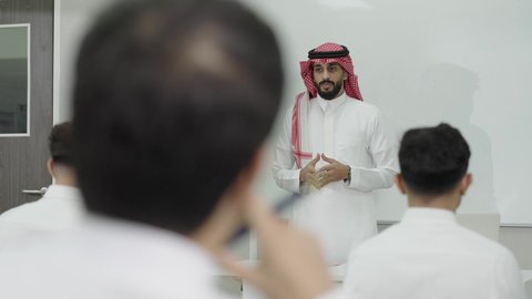 التعليم العالي في الجامعات والكليات الأكاديمية, الجلوس في الفصل الدراسي، اختبارات وكتابة التقارير والأبحاث العلمية، أستاذ جامعي عربي خليجي سعودي يرتدي الثوب التقليدي والشماغ يقوم بشرح المعلومات على الطلاب، جودة التعليم الجامعي في المملكة العربية السعودية