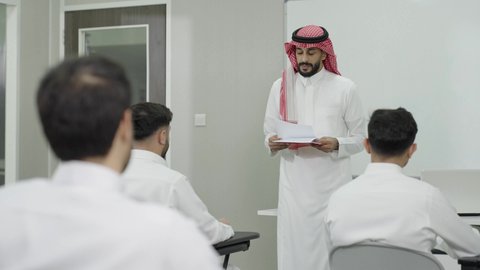 الجلوس في الفصل الدراسي، التعليم العالي في الجامعات والكليات الأكاديمية, اختبارات وكتابة التقارير والأبحاث العلمية، أستاذ جامعي عربي خليجي سعودي يرتدي الثوب التقليدي والشماغ يقوم بتوزيع الأوراق على الطلاب، جودة التعليم الجامعي في المملكة العربية السعودية