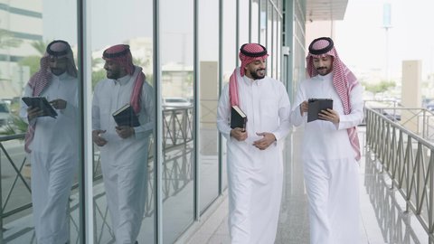 الحضور للدوام في الوقت المحدد، التعاون بين زملاء العمل، المناقشة في المناهج والدروس الجامعية، استخدام جهاز تابلت حديث ومتطور، إنجاز مهام العمل، أستاذ جامعي عربي خليجي سعودي يرتدي الشماغ والثوب التقليدي يمشي مع صديقه ويمسك بيده كتاب