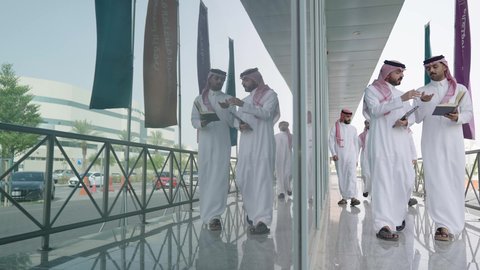 التعاون بين زملاء العمل، المناقشة في المناهج والدروس الجامعية، استخدام جهاز تابلت حديث ومتطور، إنجاز مهام العمل، أستاذ جامعي عربي خليجي سعودي يرتدي الشماغ والثوب التقليدي يمشي مع صديقه ويمسك بيده كتاب، الحضور للدوام في الوقت المحدد في الشركة