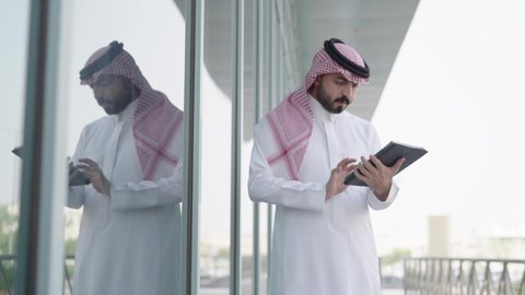 The use of a modern and advanced technical device, accomplishing remote work tasks, an Arab Gulf Saudi man wearing a traditional headscarf and thobe holding a mobile tablet in his hand, attending work on time at the company.