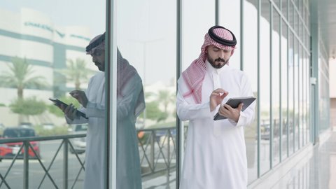 إنجاز مهام العمل عن بعد، رجل عربي خليجي سعودي يرتدي الشماغ والثوب التقليدي يمسك بيده التابلت المحمول، جهاز تقني حديث ومتطور، الحضور للدوام في الوقت المحدد في الشركة