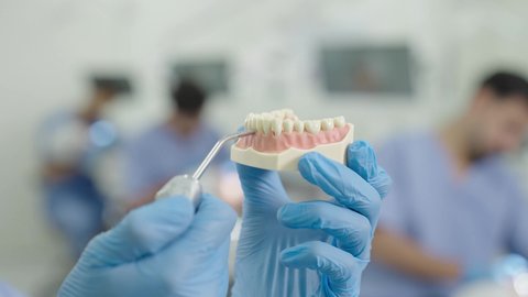 تصوير مقرب ليد طالب جامعي سعودي بكلية الأسنان يرتدي القفازات يقوم بفحص مجسم طاقم أسنان، مفهوم التدريب العملي، استخدام معدات وأدوات طب الأسنان، الأساليب العلاجية المتبعة للعناية بالفم، معالجة التسوس وتركيب الحشوات