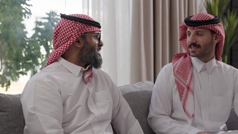 التسامر وتبادل الاحاديث، جلسة عائلية سعيدة، تعابير المودة والمحبة، الاستمتاع بقضاء وقت مع الابناء، رجل كبير بالسن عربي خليجي سعودي يرتدي الثوب التقليدي والشماغ يجلس على الاريكة بجانب ابنه الشاب في غرفة الجلوس