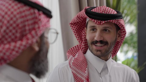 التسامر وتبادل الاحاديث، جلسة عائلية سعيدة، تعابير المودة والمحبة، الاستمتاع بقضاء وقت مع الابناء، رجل كبير بالسن عربي خليجي سعودي يرتدي الثوب التقليدي والشماغ يجلس على الاريكة بجانب ابنه الشاب في غرفة الجلوس