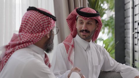 التسامر وتبادل الاحاديث، جلسة عائلية سعيدة، تعابير المودة والمحبة، الاستمتاع بقضاء وقت مع الابناء، رجل كبير بالسن عربي خليجي سعودي يرتدي الثوب التقليدي والشماغ يجلس على الاريكة بجانب ابنه الشاب في غرفة الجلوس