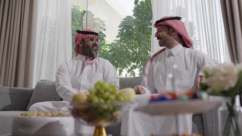  الاستمتاع بقضاء وقت مع الابناء، رجل كبير بالسن عربي خليجي سعودي يرتدي الثوب التقليدي والشماغ يجلس على الاريكة بجانب ابنه الشاب في غرفة الجلوس، التسامر وتبادل الاحاديث، جلسة عائلية سعيدة، تعابير المودة والمحبة