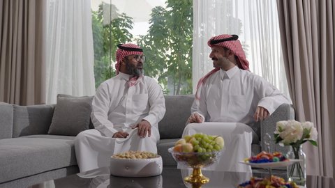  الاستمتاع بقضاء وقت مع الابناء، رجل كبير بالسن عربي خليجي سعودي يرتدي الثوب التقليدي والشماغ يجلس على الاريكة بجانب ابنه الشاب في غرفة الجلوس، التسامر وتبادل الاحاديث، جلسة عائلية سعيدة، تعابير المودة والمحبة