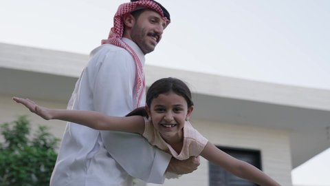 Spending quality time with family and children, a Saudi Arabian Gulf man wearing a traditional thobe and shemagh lifts his daughter high with happy gestures, maintaining family ties, taking care of children, playing, having fun and entertaining with the family outdoors