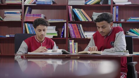 إنجاز الواجبات اليومية، طالبان عربيان سعوديان خليجييان يرتديان الزي المدرسي يجلسان في المكتبة المدرسية، الاستمتاع بقراءة الكتب المدرسية، التعليم في المملكة العربية السعودية
