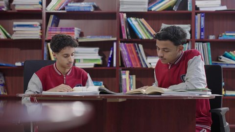 Performing school assignments, two Arab Saudi Gulf students wearing school uniforms are sitting in the school library, enjoying reading school books, education in the Kingdom of Saudi Arabia.