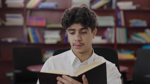 The interest in browsing useful books, love for culture and exploration, a close-up shot of a Saudi Gulf Arab student wearing a white thobe and a shemagh reading a book, spending free time learning and self-educating, visiting the school library, education and studying in Saudi Arabia, developing reading skills for students.