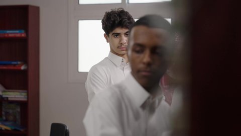 Cooperative learning among peers, discussing study materials, a Saudi Gulf Arab student wearing a white thobe and a shemagh holding a book and talking to his classmate in the school library.