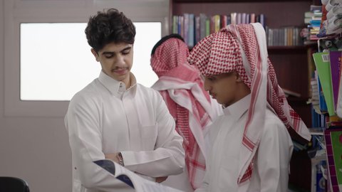 Studying subjects collectively, discussing academic materials, a Saudi Gulf Arab student wearing a white thobe and a ghutrah is holding a book in his hand and talking to his classmate in the school library.