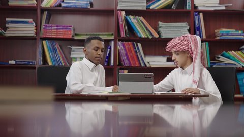 Two Saudi Arabian Gulf Arab students are sitting in front of a tablet in the school library, the concept of self-education, the school library, an advanced educational school environment, academic schools in the Kingdom of Saudi Arabia, discussing the study materials.