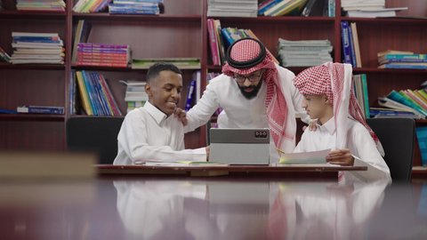 موسم العودة إلى المدارس, أهداف التربية والتعليم في المملكة العربية السعودية، طالبان عربيان خليجيان سعوديان يرتديان الثوب الابيض يجلسان داخل مكتبه المدرسة برفقة معلمهم، استخدام التابلت فى الدراسة، تعابير السعادة و السرور