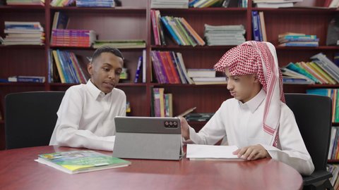 مناقشة المواد الدراسيه، التعليم والدراسة في السعودية، طالبان عربيان خليجيان سعوديان يرتديان الثوب الابيض يجلسان داخل مكتبه المدرسة، استخدام التابلت فى الدراسة