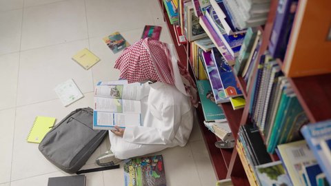 A developed educational school environment, preparation for school exams, a Saudi Gulf Arab student wearing a white thobe and a shemagh sitting next to the library reading a book, studying and reviewing, academic schools in the Kingdom of Saudi Arabia.
