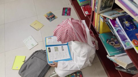 Academic schools in the Kingdom of Saudi Arabia, preparing for school exams, a Gulf Arab Saudi student wearing a white thobe and a shemagh sitting next to the library reading a book, studying and reviewing, an advanced educational school environment.