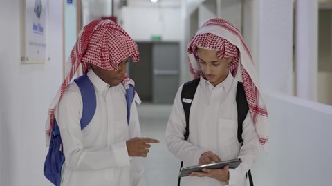 التعاون الدراسي والمشاركة في حل التمارين والواجبات، مفهوم التعليم الإلكتروني، مناقشة المواد الدراسيه، طالب عربي خليجي سعودي يرتدي الثوب الابيض و الشماغ و الحقيبة المدرسية يقف برفقة زميله ، استخدام الجهاز اللوحي فى الدراسة ، استخدام الاجهزة التقنية الحديثة