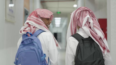 ايماءات السعادة و السرور،تعابير السلام و التحية،  العودة للمدارس في المملكة العربية السعودية، الاستعداد للعام الدراسي الجديد, متابعة الدروس والواجبات،تصوير من الخلف لطالبان عربيان خليجيان سعوديان يرتديان الثوب الابيض و الشماغ و الحقيبة المدرسية يسيران داخل ممر المدرسة