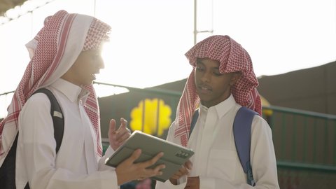 العمل الجماعي بين الطلاب أثناء المذاكرة، مفهوم التعليم الإلكتروني، مناقشة المواد الدراسيه، طالب عربي خليجي سعودي يرتدي الثوب الابيض و الشماغ و الحقيبة المدرسية يقف برفقة زميله ، استخدام الجهاز اللوحي فى الدراسة ، استخدام الاجهزة التقنية الحديثة