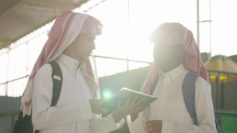 تأدية الواجبات المدرسية، مفهوم التعليم الإلكتروني، مناقشة المواد الدراسيه، طالب عربي خليجي سعودي يرتدي الثوب الابيض و الشماغ و الحقيبة المدرسية يقف برفقة زميله ، استخدام الجهاز اللوحي فى الدراسة ، استخدام الاجهزة التقنية الحديثة