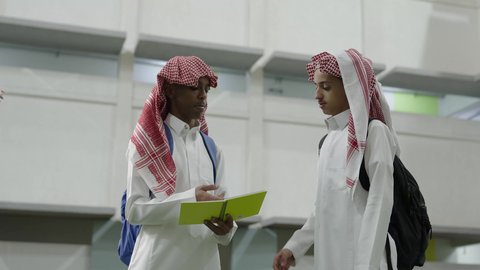 التفاعل الجماعي لمناقشة الدروس والواجبات المدرسية، التعليم في المملكة العربية السعودية، طالبان عربيان خليجيان سعوديان يرتديان الثوب الابيض و الشماغ يحملان الحقيبة المدرسية يقفان داخل المدرسة ، الاستعداد للبدء بالعام الدراسي الجديد، العودة الى المدارس التعليمية في المملكة