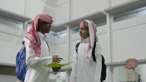Exchange of opinions about lessons and study content among students, teamwork and cooperation among colleagues, education in the Kingdom of Saudi Arabia, two Arab Gulf Saudi students wearing white thobes and shemaghs carrying school bags standing inside the school, preparing to start the new school year, returning to educational schools in the Kingdom.