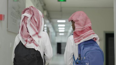 The importance of education in building the future, the return to schools in the Kingdom of Saudi Arabia, preparation for the new school year, following lessons and assignments, a shot from behind of two Arab Gulf Saudi students wearing the white thobe and the shemagh, carrying school bags, walking inside the school corridor.