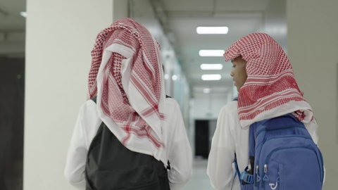 العودة للمدارس في المملكة العربية السعودية، الاستعداد للعام الدراسي الجديد، أهمية التعليم في بناء المستقبل, متابعة الدروس والواجبات،تصوير من الخلف لطالبان عربيان خليجيان سعوديان يرتديان الثوب الابيض و الشماغ و الحقيبة  المدرسية يسيران داخل ممر المدرسة