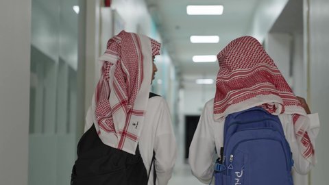الاستعداد للعام الدراسي الجديد، العودة للمدارس في المملكة العربية السعودية، أهمية التعليم في بناء المستقبل, متابعة الدروس والواجبات،تصوير من الخلف لطالبان عربيان خليجيان سعوديان يرتديان الثوب الابيض و الشماغ و الحقيبة  المدرسية يسيران داخل ممر المدرسة