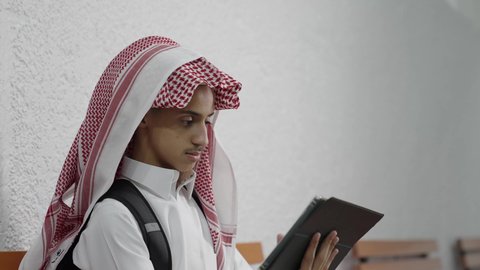 The concept of e-learning, teamwork and collaboration among colleagues, discussion of study materials, a Saudi Gulf Arab student wearing a white thobe and a shemagh with a school bag sitting with a colleague, using a tablet in studying, using modern technological devices.