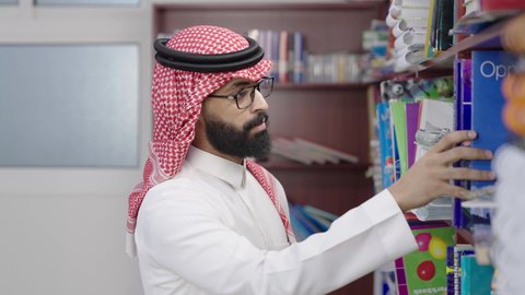 ممارسة الهوايات المفضلة ، قضاء وقت ممتع ، معلم عربي خليجي سعودي يرتدي الثوب الابيض و الشماغ يقف بجوار مكتبة خشبية في المكتبة المدرسية، الاستمتاع بقراءة الكتب المدرسية، التعليم في المملكة العربية السعودية